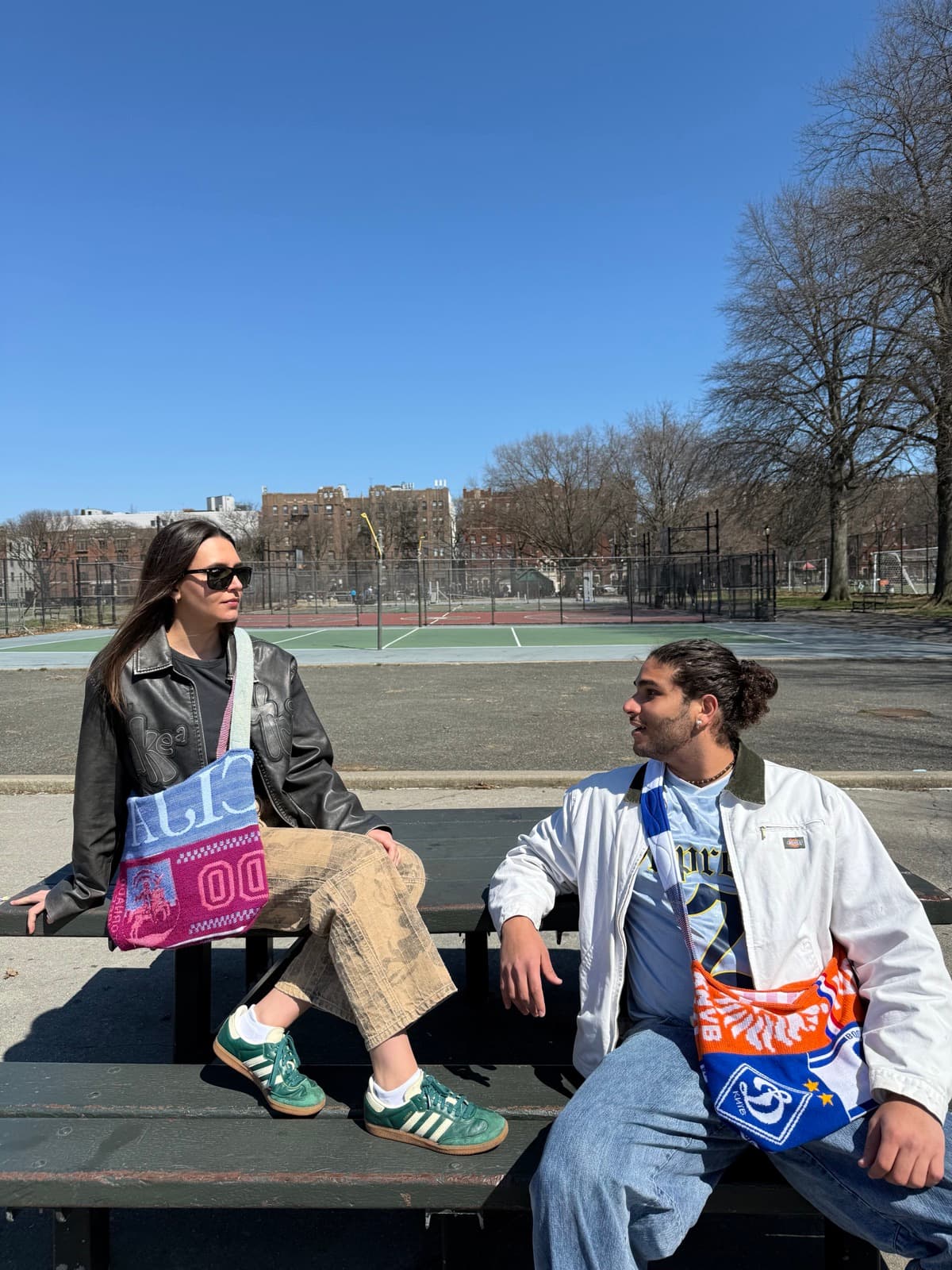 Two friends on a park bench carrying Extra Time scarf bags, ball courts behind them